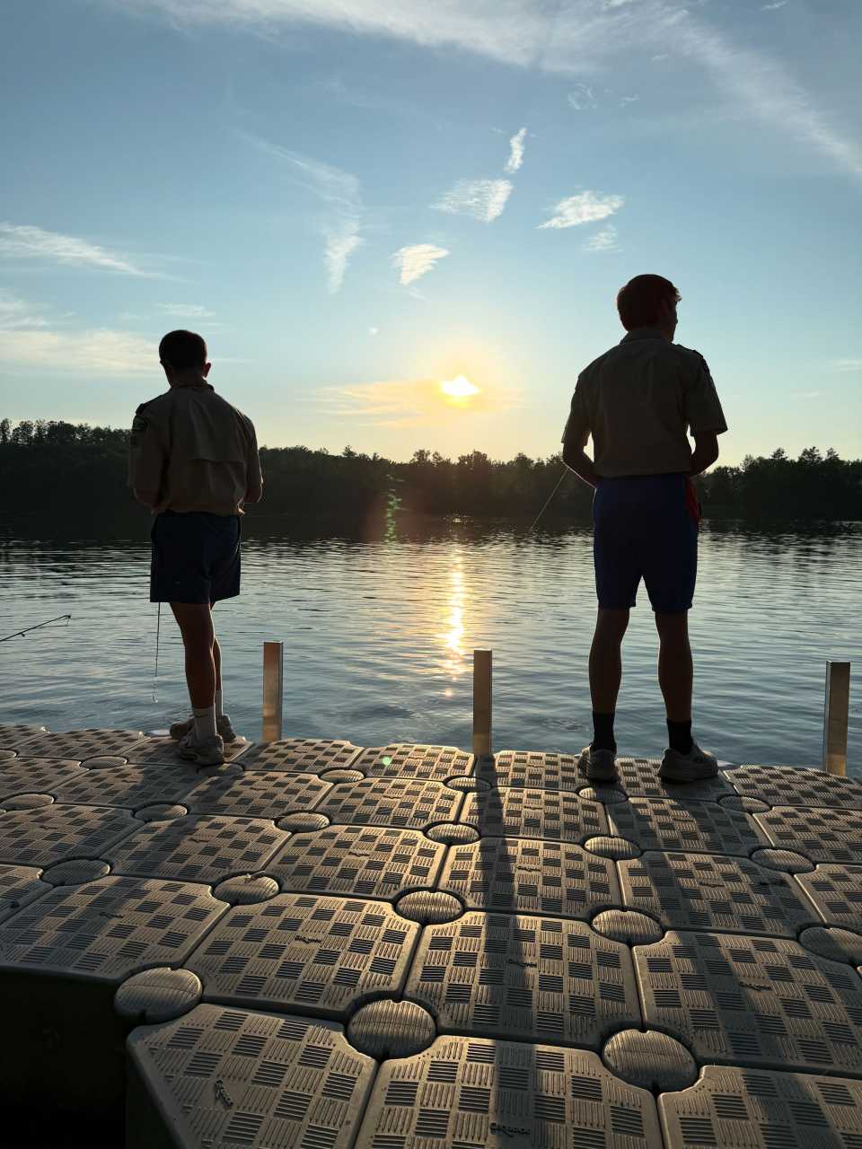 The sun sets on the horizon behind the lake, with the silhouette of two fishing scouts standing on the pier in the foreground